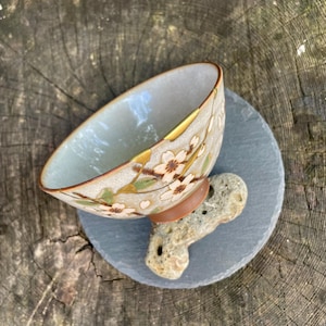 Può includere: Ciotola in ceramica decorativa con un motivo floreale nei toni del beige, marrone e oro. La ciotola poggia su un sottobicchiere di pietra grigia ed è sostenuta da una piccola roccia beige porosa. L'interno della ciotola è di un verde-azzurro chiaro.