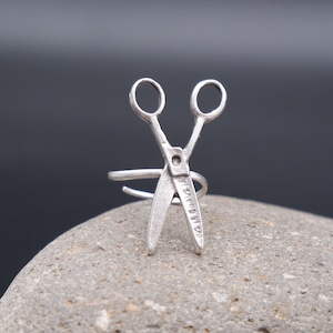 May include: A silver ring with a detailed design of a pair of scissors. The ring is sitting on a gray rock.