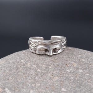 May include: A silver-toned ring with a sculpted face design. The ring features a detailed depiction of a face with closed eyes and flowing hair. The ring is set on a gray stone against a dark background, highlighting the intricate details of the jewelry.