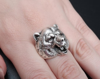 May include: A silver ring featuring a detailed bear head design. The bear's mouth is open, showing its teeth. The ring is worn on a person's finger.