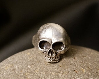 May include: A silver skull ring with detailed features. The ring is sitting on a gray stone.