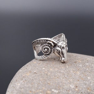 Może przedstawiać: Srebrny pierścionek z detalicznym wzorem głowy barana. Rogi barana są zakrzywione, a głowa skierowana do przodu. Pierścionek leży na szarej powierzchni kamiennej.