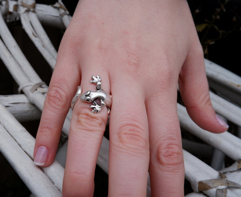 May include: A silver ring shaped like a gecko lizard. The ring is worn on a person's finger.