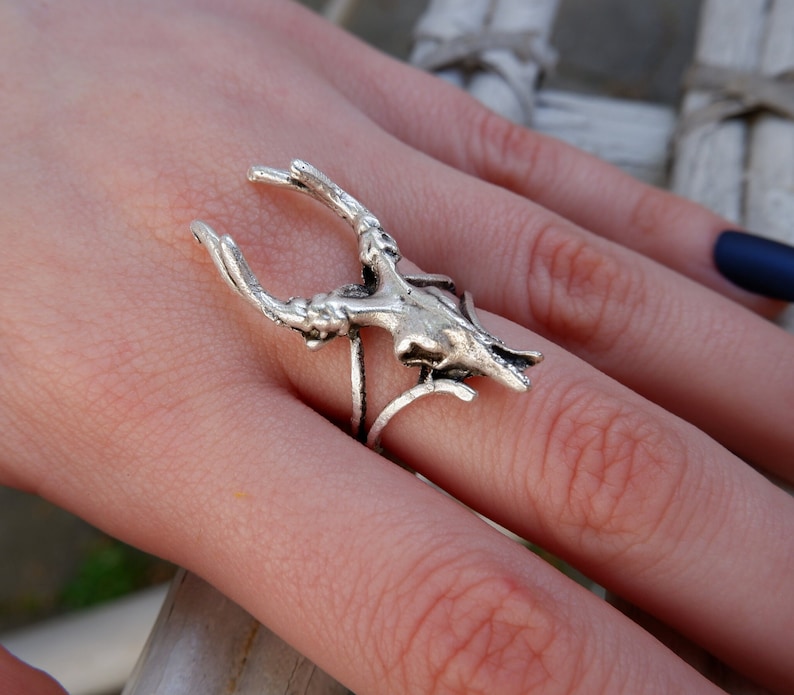 May include: A silver ring featuring a detailed deer skull design with antlers. The ring is worn on a finger.