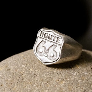May include: A silver ring with a Route 66 shield design. The ring is on a gray rock.