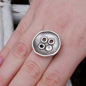 May include: A silver ring featuring a large button-shaped design. The button has five holes in it.