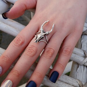 May include: A silver ring with a deer skull design. The ring is worn on a finger with dark blue nail polish.