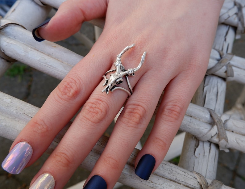 May include: A silver ring with a deer skull design. The ring is worn on a finger with dark blue nail polish.