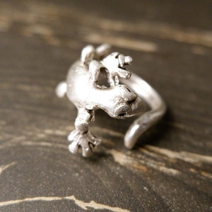 May include: A silver ring featuring a detailed gecko design. The gecko is positioned on its back with its legs outstretched. The ring is open and adjustable.