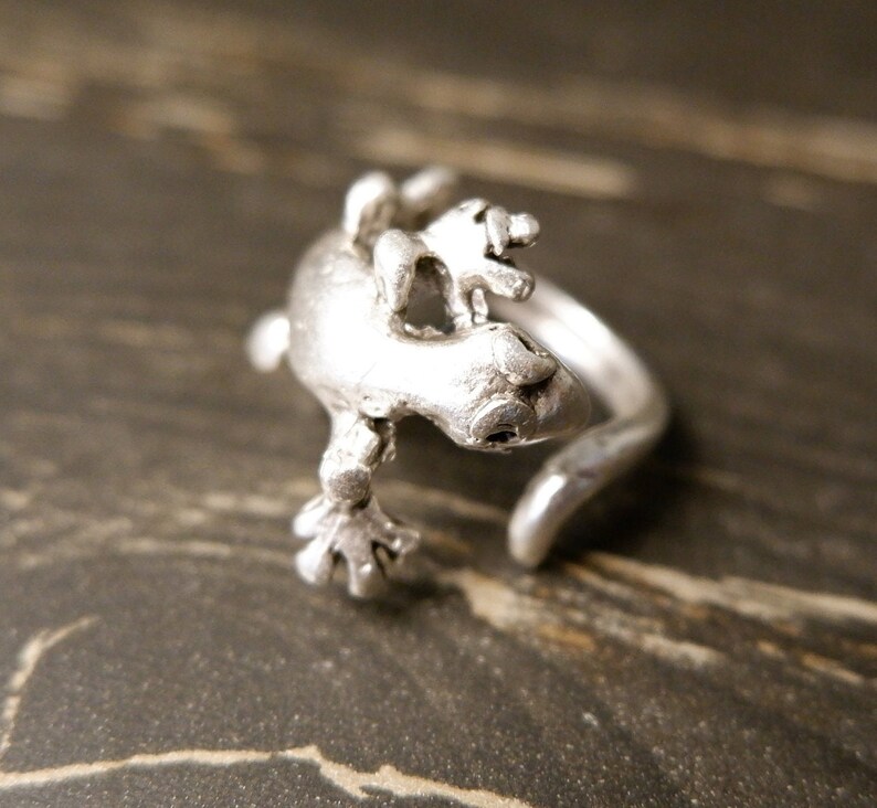 May include: A silver ring featuring a detailed gecko design. The gecko is positioned on its back with its legs outstretched. The ring is open and adjustable.