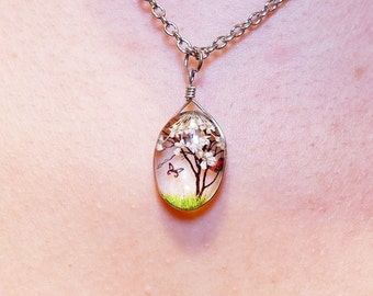 Puede incluir: Un collar de cadena plateada con un colgante ovalado transparente que contiene un pequeño árbol de flores blancas con dos mariposas y una mariquita roja.