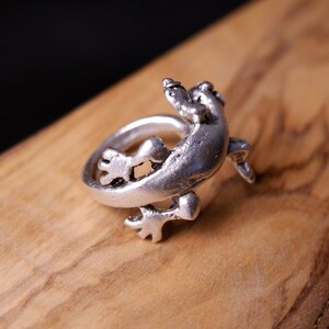 May include: A silver ring featuring a detailed lizard design. The lizard is depicted in a curled position, with its tail wrapping around the ring band.