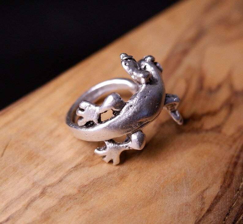 May include: A silver ring featuring a detailed lizard design. The lizard is depicted in a curled position, with its tail wrapping around the ring band.