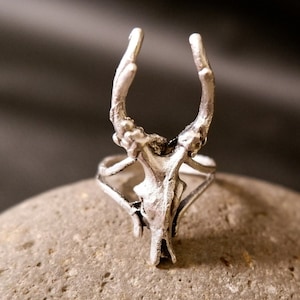 May include: A silver ring with a deer skull design. The antlers are curved upwards and the skull is detailed.