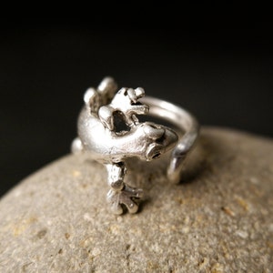 May include: A silver ring featuring a detailed gecko design. The gecko is perched on a silver band, with its legs and tail curled around the band.
