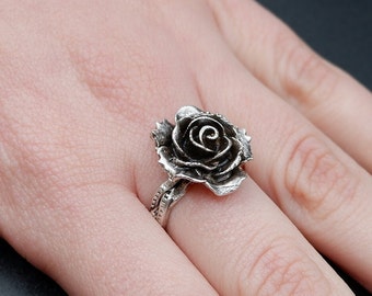 Könnte beinhalten: Ein silberner Ring mit einem detaillierten Rosenmotiv. Die Rose ist dreidimensional und hat eine dunkelgraue Oberfläche.