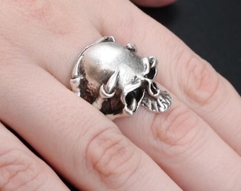 May include: A silver skull ring with detailed facial features. The ring is worn on a finger, showcasing the skull's intricate design. The skull's eye sockets, nose, and teeth are clearly visible, giving it a realistic appearance. The ring is a gothic or biker-style accessory.