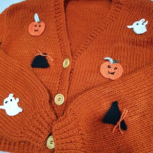 May include: An orange, knitted cardigan sweater with Halloween-themed appliqu&eacute;s. The sweater features pumpkin, ghost, and witch hat designs. It has a V-neck, wooden buttons, and a cozy, textured appearance, perfect for the fall season.