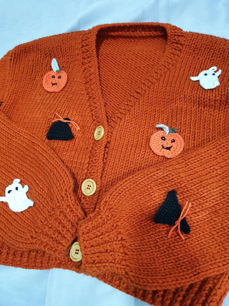 May include: An orange, knitted cardigan sweater with Halloween-themed appliqu&eacute;s. The sweater features pumpkin, ghost, and witch hat designs. It has a V-neck, wooden buttons, and a cozy, textured appearance, perfect for the fall season.