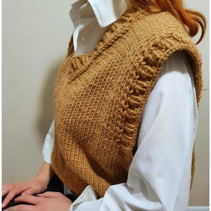 Puede incluir: Un chaleco tejido a mano de color mostaza con un diseño de cuello en V. El chaleco se lleva sobre una camisa blanca con cuello. La textura tejida es visible y los bordes tienen una puntada decorativa.
