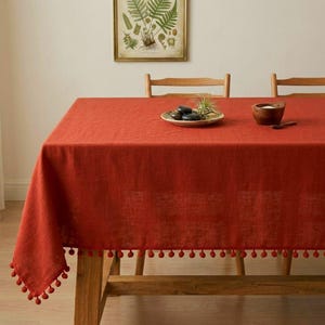 Può includere: Una tovaglia rettangolare color ruggine con finiture a pompon. La tovaglia è drappeggiata su un tavolo di legno. Una piccola ciotola di legno, un piatto con sassi e una piccola pianta sono sul tavolo. Una stampa botanica incorniciata è appesa al muro.