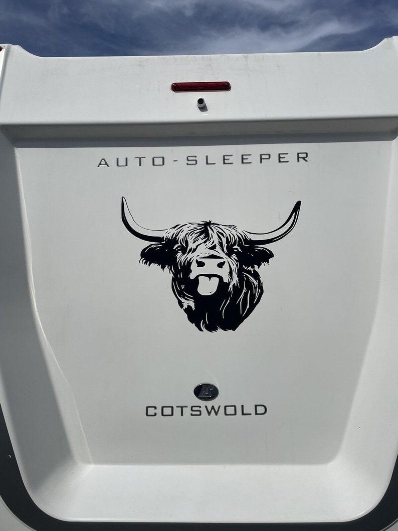 Pu&ograve; includere: Un camper bianco con le parole "AUTO-SLEEPER" e "COTSWOLD" in lettere nere. Un'illustrazione in bianco e nero di una mucca delle Highlands con la lingua di fuori &egrave; centrata sul camper.
