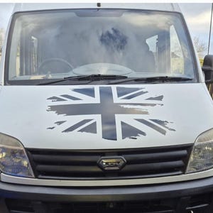 Könnte beinhalten: Ein weißer Van mit einem schwarz-weißen Union-Jack-Flaggenaufkleber auf der Motorhaube. Der Van hat einen schwarzen Kühlergrill und Scheinwerfer.
