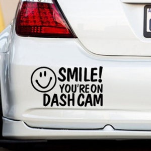 Puede incluir: Un coche blanco con una pegatina negra en el parachoques que dice "¡Sonríe! Estás en Dash Cam" con un icono de cara sonriente.