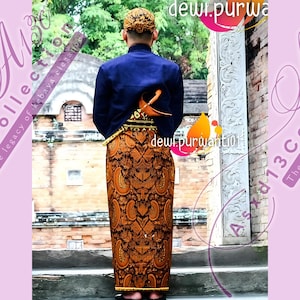 Könnte beinhalten: Ein Mann trägt ein blaues Hemd und einen traditionellen indonesischen Sarong mit gold-schwarzem Muster. Der Sarong ist in der Taille gebunden und hat einen dekorativen Knoten vorne. Der Mann steht vor einer Steinmauer mit einem geschnitzten Design.