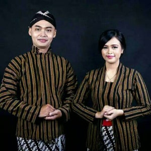 Könnte beinhalten: Ein Mann und eine Frau in traditioneller indonesischer Kleidung. Der Mann trägt ein schwarz-gold gestreiftes Hemd mit einem schwarzen Hut. Die Frau trägt eine schwarz-gold gestreifte Jacke mit einem schwarz-weiß gemusterten Rock.