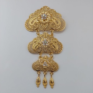 Peut inclure: Une broche en filigrane dorée à trois niveaux de motifs complexes et de strass transparents. Le niveau inférieur comporte trois pendentifs en forme de goutte.