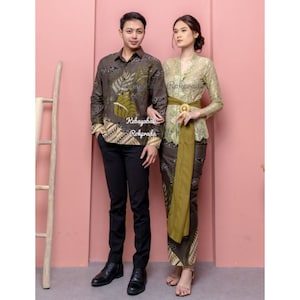 May include: A couple wearing matching brown and green patterned clothing. The man is wearing a long-sleeved shirt with a leaf design and black pants. The woman is wearing a lace top with a green sash and a matching skirt. The text "Kebayabali Rokprada" is visible on the shirt.