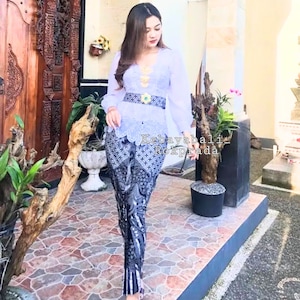 Peut inclure: Une femme portant une kebaya bleu clair avec une bordure en dentelle blanche et une jupe à motifs noir et blanc. Elle porte une ceinture dorée avec un bijou vert. Le texte "Kebaya Bali Rok Prada" est visible sur l'image.