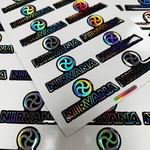 May include: A close-up of a stack of holographic stickers with the word "NIRVANA" printed in black letters. The stickers feature a rainbow-colored swirl design.