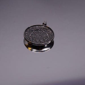 Pu&ograve; includere: Un ciondolo circolare in metallo color argento con intricati dettagli in rilievo sulla superficie. Il design presenta un motivo geometrico centrale circondato da un bordo di piccoli simboli ripetuti. Un piccolo occhiello &egrave; fissato al bordo superiore.