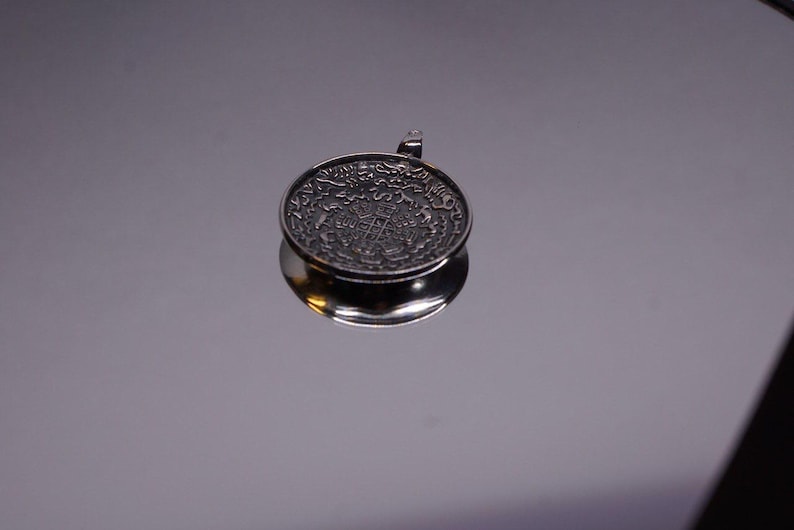 Pu&ograve; includere: Un ciondolo circolare in metallo color argento con intricati dettagli in rilievo sulla superficie. Il design presenta un motivo geometrico centrale circondato da un bordo di piccoli simboli ripetuti. Un piccolo occhiello &egrave; fissato al bordo superiore.