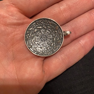 Pu&ograve; includere: Un ciondolo circolare color argento caratterizzato da un intricato design in rilievo con un motivo a griglia centrale circondato da varie figure animali e simboli astratti. La superficie metallica presenta una finitura ossidata scura.