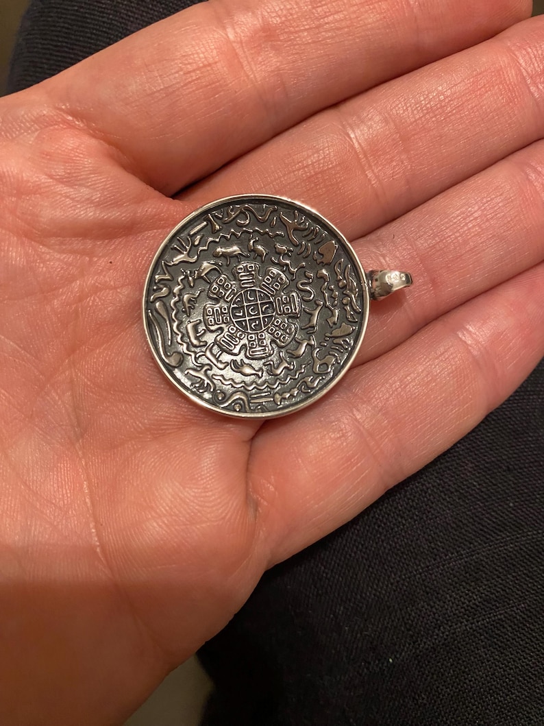 Pu&ograve; includere: Un ciondolo circolare color argento caratterizzato da un intricato design in rilievo con un motivo a griglia centrale circondato da varie figure animali e simboli astratti. La superficie metallica presenta una finitura ossidata scura.