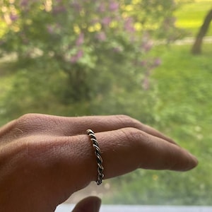 Puede incluir: Un anillo plateado con un diseño de cuerda trenzada. El anillo se lleva en un dedo, mostrando su intrincada textura y brillo metálico. El fondo es una vista borrosa de un césped verde y flores moradas.