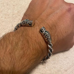 以下が含まれることがあります： ねじれた金属バンドが特徴のシルバー製カフブレスレット。両端には詳細な動物の頭のモチーフがあしらわれています。それぞれの動物の頭の側面には、小さな円形の緑色の宝石がはめ込まれています。