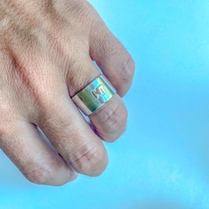 Op de afbeelding: Een brede zilveren ring met een gepolijste metalen afwerking en de letters NT gegraveerd in een contrasterende goudtint op het vooroppervlak. De ring heeft een glad, plat profiel en zit horizontaal om de vinger.