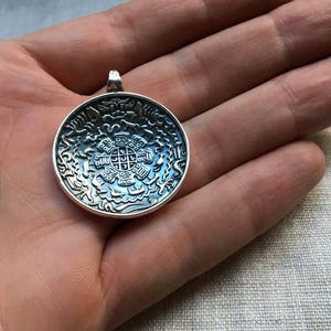 Pu&ograve; includere: Un ciondolo rotondo in argento con un intricato design in rilievo che presenta una figura geometrica centrale circondata da motivi stilizzati di animali e onde. Il metallo ha una finitura lucida con aree incassate scure.