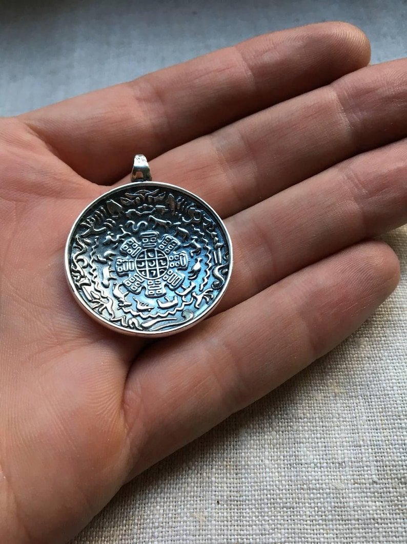 Pu&ograve; includere: Un ciondolo rotondo in argento con un intricato design in rilievo che presenta una figura geometrica centrale circondata da motivi stilizzati di animali e onde. Il metallo ha una finitura lucida con aree incassate scure.