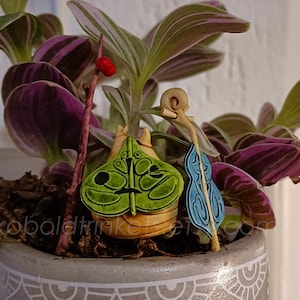 Peut inclure: Une figurine verte et brune avec une feuille bleue et blanche, assise dans un pot avec une plante violette et verte. Le pot a un motif décoratif.