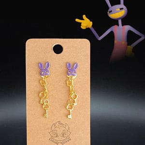 Puede incluir: Pendientes de conejito morados con dijes de llaves doradas. Se exhiben en una tarjeta marrón con el texto "Kobold Trinkets". Un personaje de dibujos animados morado con guantes amarillos está en el fondo.