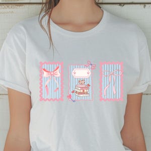 May include: White t-shirt with three rectangular designs. Each design features pink bows, vertical blue stripes, and a stack of books with a heart and a small cat. The designs are framed in pink.