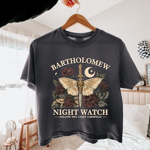 Puede incluir: Una camiseta gris carbón de manga corta con un estampado gráfico de una polilla centrada en una daga vertical, rodeada de flores oscuras y una luna creciente. El texto dice BARTHOLOMEW NIGHT WATCH y FOLLOW THE LIGHT CAREFULLY.