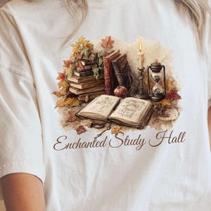 May include: Cream-colored t-shirt featuring a detailed illustration of a study scene. The design includes stacks of books, autumn leaves, a lit candle, and an hourglass. The text "Enchanted Study Hall" is printed below the illustration.
