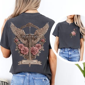 May include: Dark gray t-shirt featuring a detailed design on the back with a sword, wings, roses, and the text "VALKYRIE TRAINING CAMP." A smaller design with a sword and rose is on the front. The shirt is a comfortable, casual style.