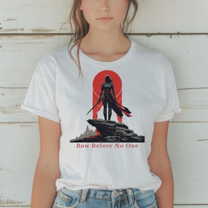 Puede incluir: Camiseta blanca con un diseño de guerrero en negro y rojo. El diseño muestra una figura con una espada sobre una formación rocosa, sobre un fondo rojo. El texto "Bow Before No One" está impreso debajo.